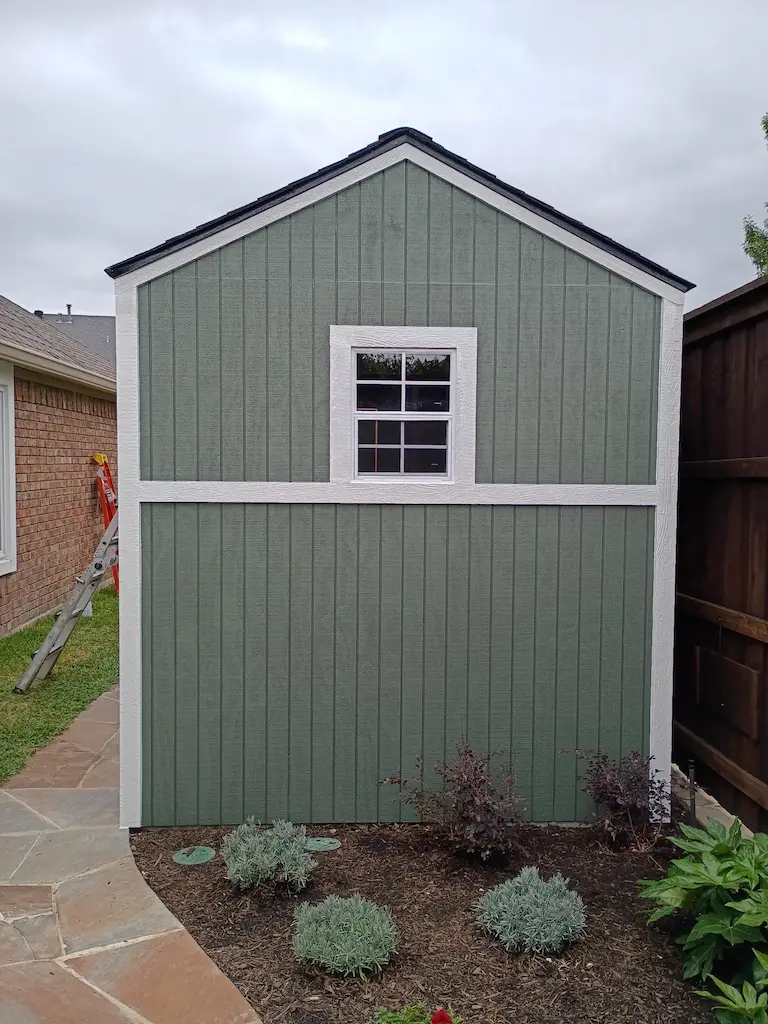 A green shed with white trim and a window is centered between a brick house and a wooden fence. A small garden with shrubs is in the foreground.