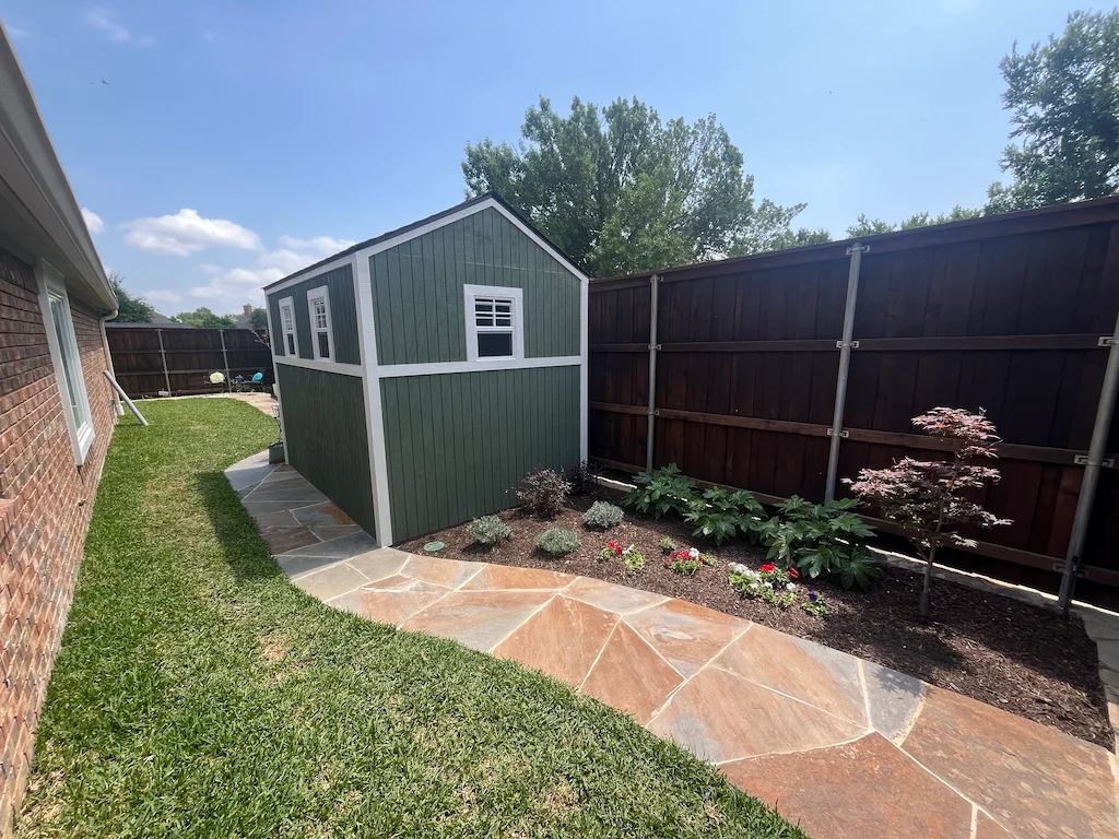 A green shed with white trim is in a backyard, surrounded by grass and a wooden fence. The shed faces a flower bed with lush plants and colorful flowers. The scene is peaceful and bright under a clear blue sky.