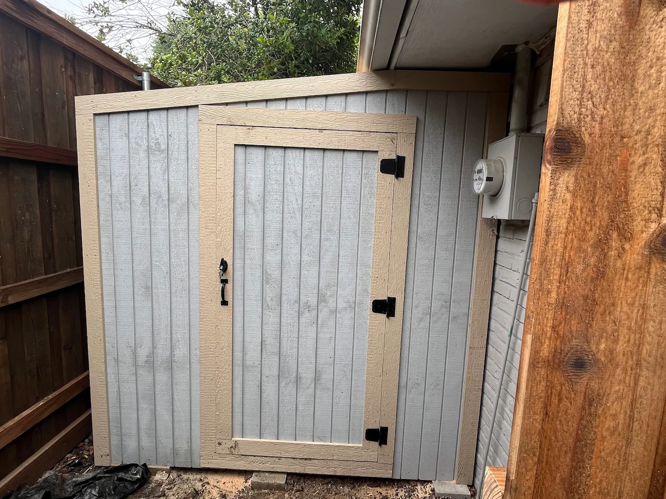 A small outdoor shed with vertical light gray panels and beige trim, set in a corner. It has a black latch, a wooden fence nearby, and a utility meter on one side.