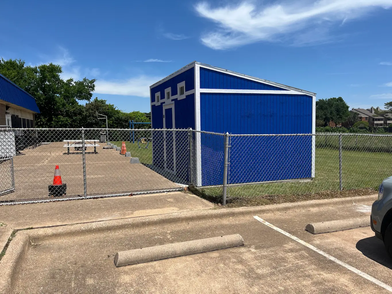 A small blue shed with white trim is enclosed by a chain-link fence. Nearby, orange cones and a picnic table sit on a concrete surface under a clear blue sky.