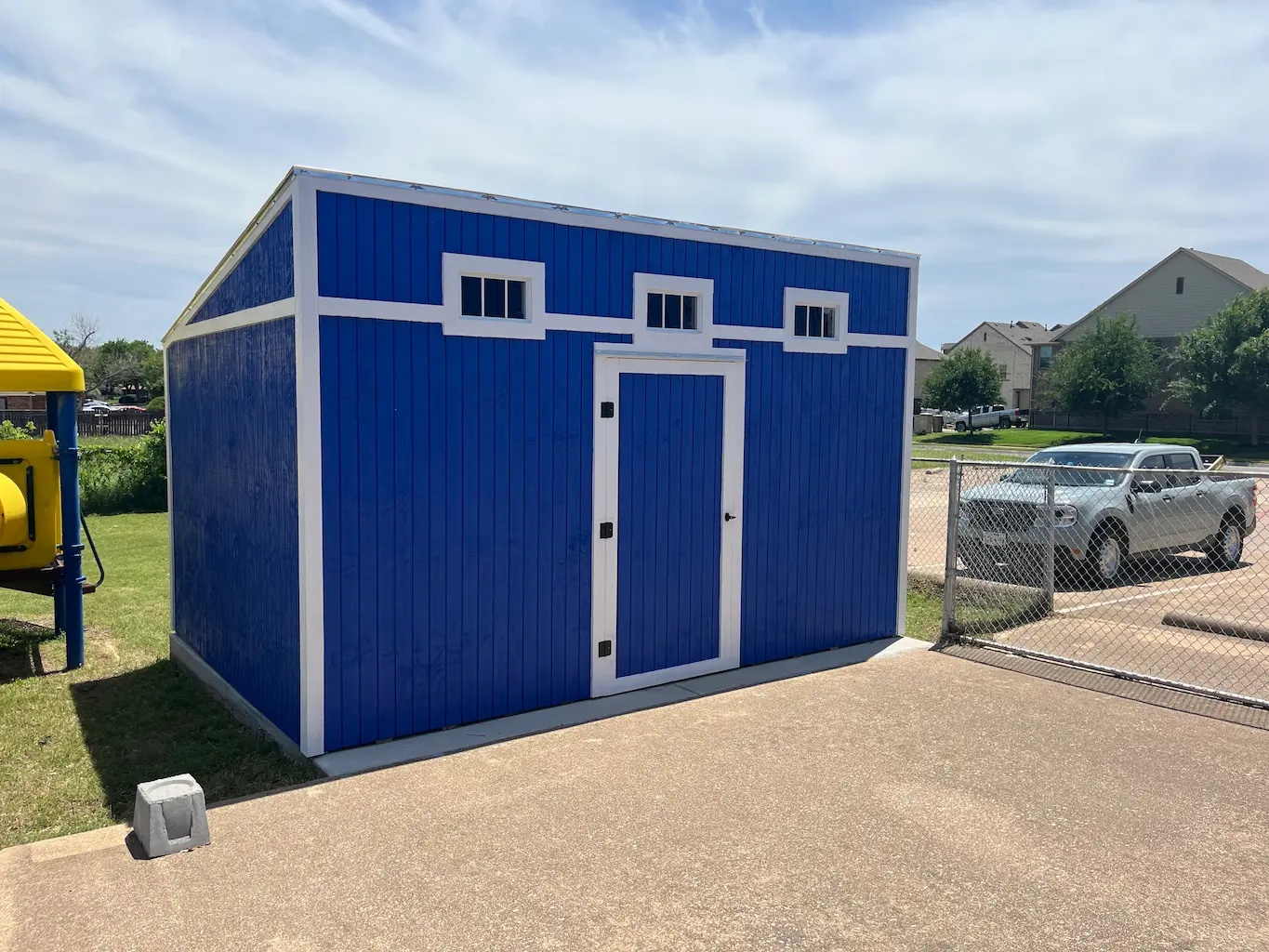 Blue shed with white trim, featuring a door and three small windows. It's set on a paved area near a chain-link fence with a parked car in the background.