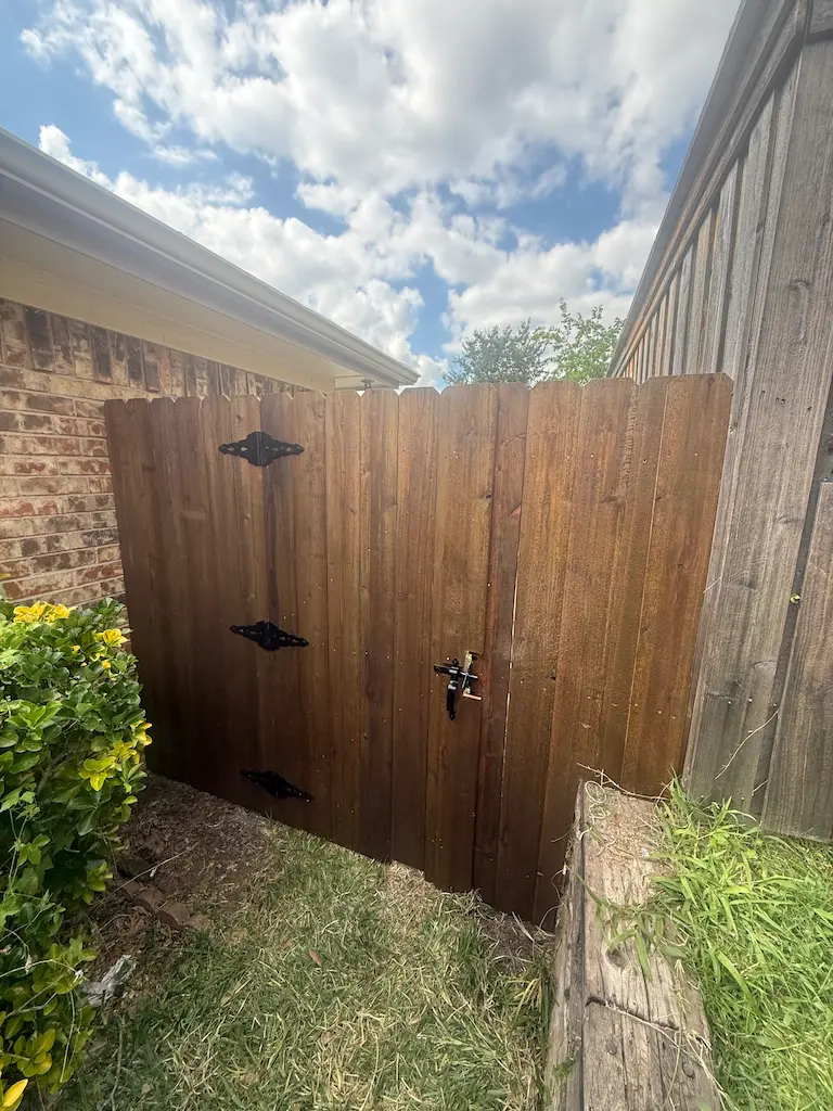 A wooden fence gate with black metal hinges stands amid brick and wood walls. Lush greenery borders the scene, under a partly cloudy blue sky.