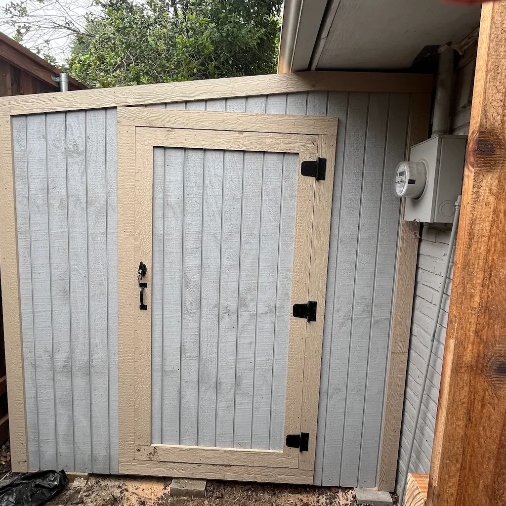 A small outdoor shed with vertical light gray panels and beige trim, set in a corner. It has a black latch, a wooden fence nearby, and a utility meter on one side.