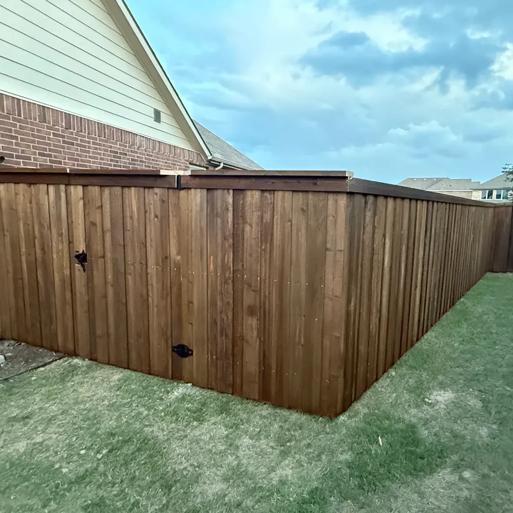 Tall wooden fence with rich brown panels encloses a yard beside a red brick house. The sky is cloudy, hinting at an approaching storm.