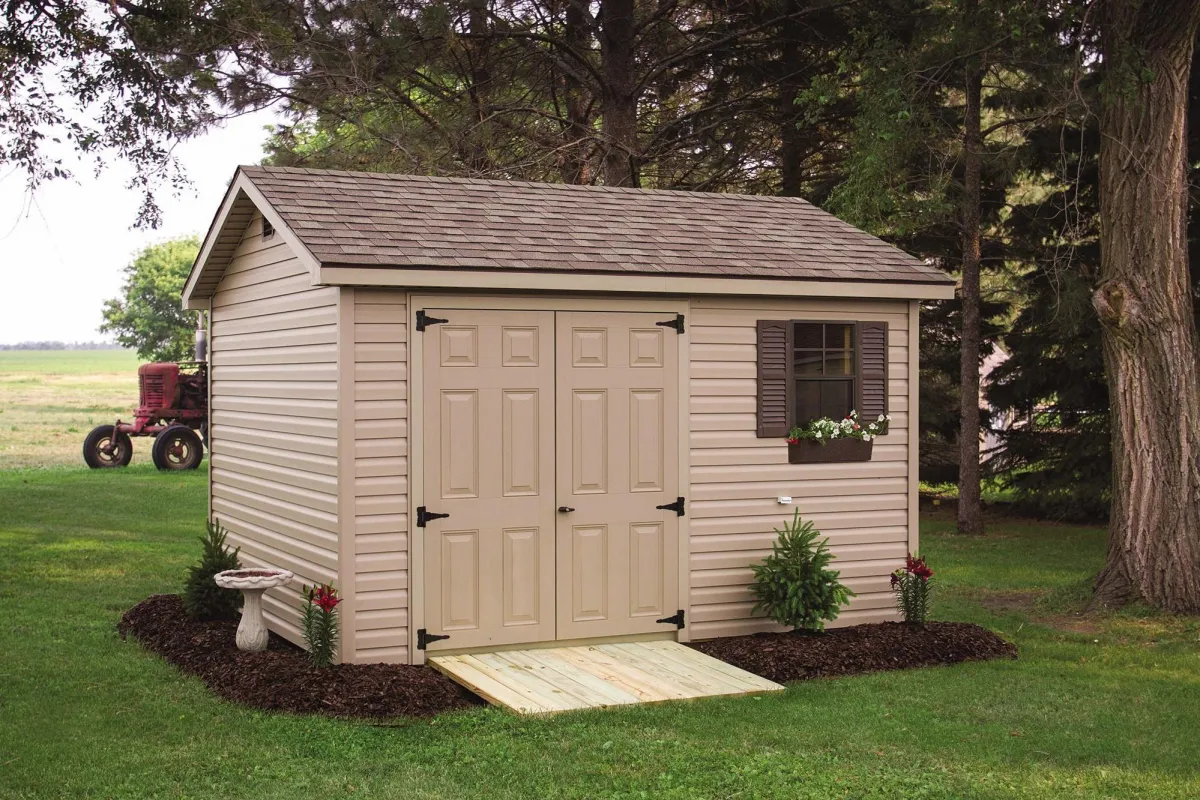 A small beige shed with double doors and a window box sits in a lush green yard, surrounded by trees. A vintage red tractor is visible in the background.