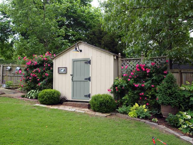 A quaint garden shed with a light green door is surrounded by lush greenery and vibrant pink flowers. The peaceful scene conveys a serene, cottage-like ambiance.